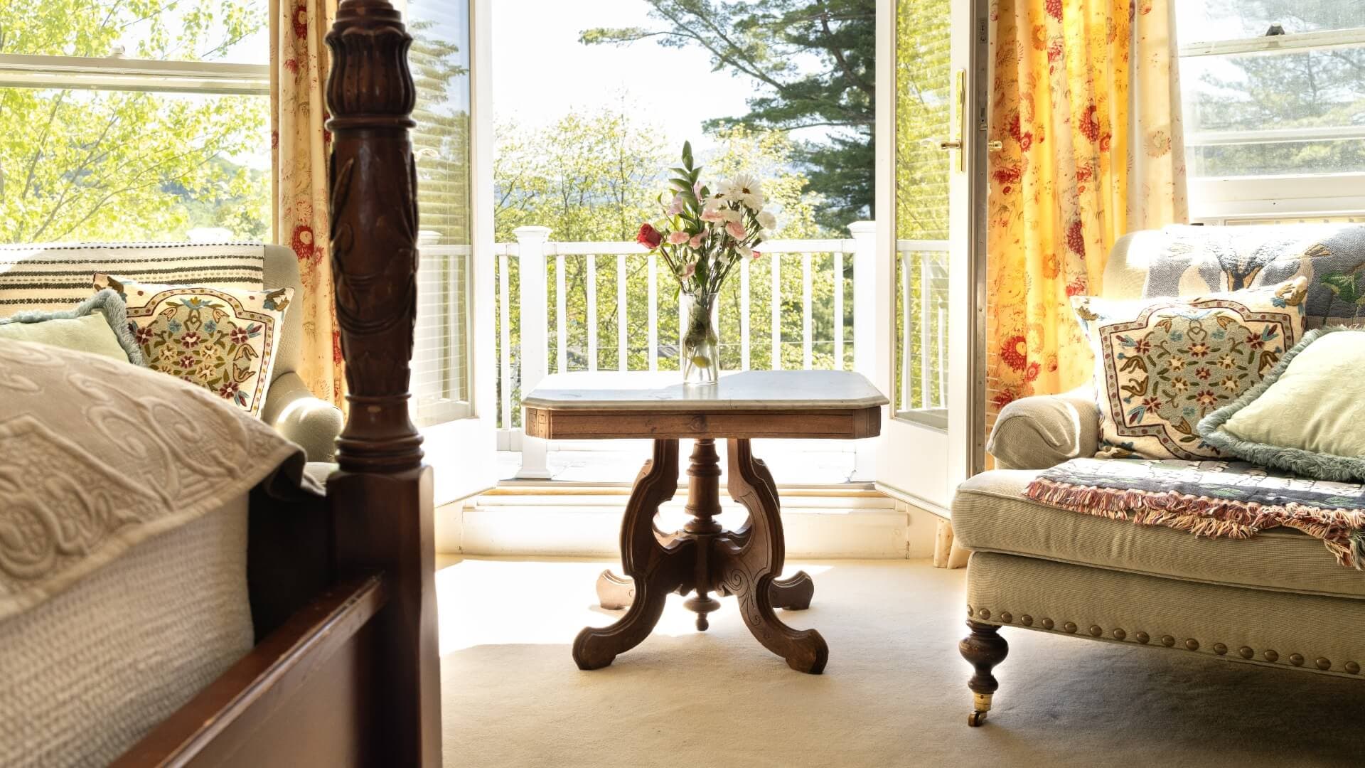 A cozy bedroom with a wooden bed, a vintage table, and a plush sofa beside large windows showcasing greenery outside.